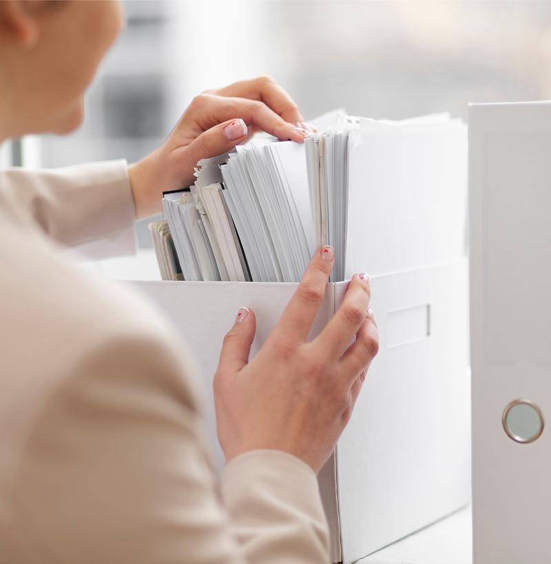 Employée de bureau cherchant un dossier spécifique dans une boîte d'archives remplie de documents imprimés.
