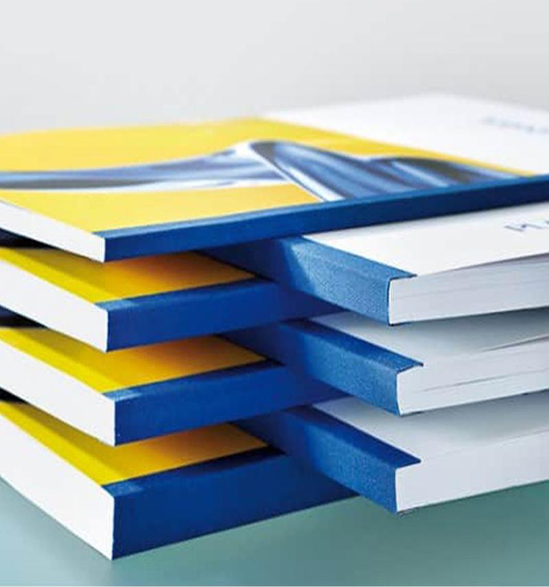 Pile de livrets d'entreprise aux couvertures jaunes et bleues, assemblés avec une reliure à dos carré collé bleu.
