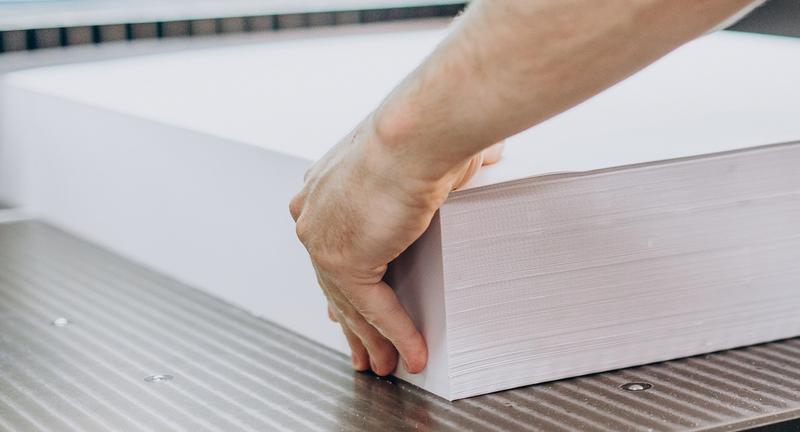 Mains d'un imprimeur ajustant une épaisse pile de feuilles de papier blanc sur une machine de découpe industrielle.